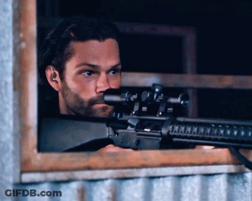 A Man With A Beard Is Holding A Rifle In Front Of A Window GIF