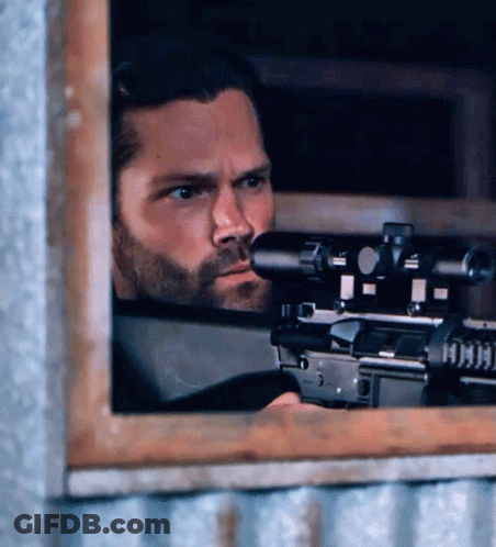 A Man With A Beard Is Holding A Sniper Rifle Behind A Window GIF