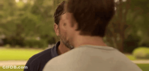A Man With A Beard Is Kissing Another Man On The Forehead In A Park . GIF