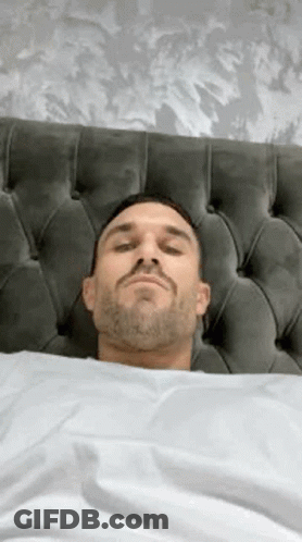 A Man With A Beard Is Laying In Bed With His Eyes Closed And His Head On The Headboard . GIF