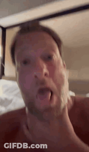 A Man With A Beard Is Laying On A Bed With His Mouth Open And His Hand On His Neck . GIF