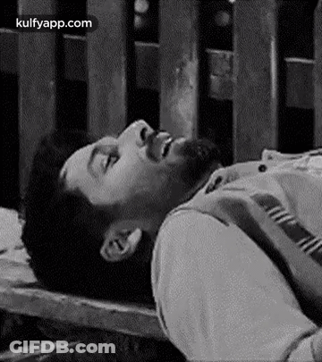 A Man With A Beard Is Laying On A Wooden Bench With His Eyes Closed . GIF