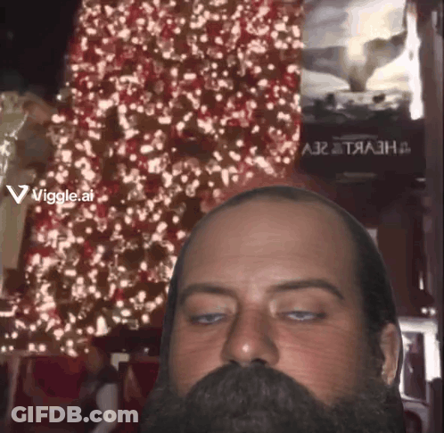 A Man With A Beard Is Looking At The Camera With A Christmas Tree In The Background And A Sign That Says Viggle.ai GIF
