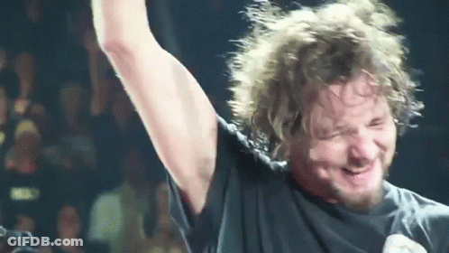 A Man With A Beard Is Singing Into A Microphone With His Arm Up In The Air GIF