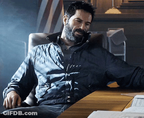 A Man With A Beard Is Sitting At A Desk Wearing A Black Jacket GIF
