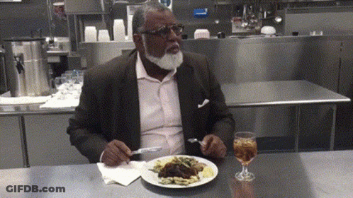 A Man With A Beard Is Sitting At A Table Eating A Plate Of Food GIF