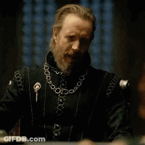 A Man With A Beard Is Sitting At A Table Wearing A Black Jacket And A Gold Chain Around His Neck . GIF
