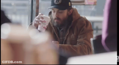 A Man With A Beard Is Sitting At A Table With A Cup In His Hand . GIF
