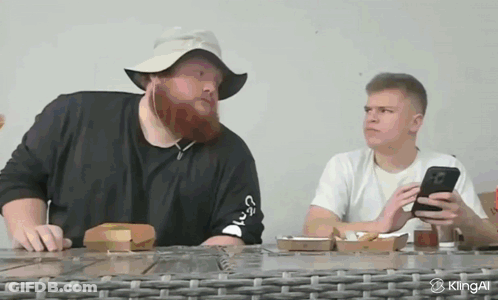 A Man With A Beard Is Sitting At A Table With A Phone In His Hand GIF