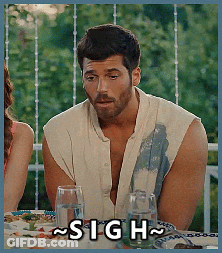 A Man With A Beard Is Sitting At A Table With A Sign That Says Sigh GIF