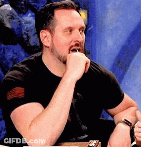 A Man With A Beard Is Sitting At A Table With His Hand On His Chin And A Watch On His Wrist . GIF