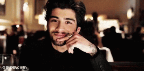 A Man With A Beard Is Sitting At A Table With His Hand On His Chin And Smiling At The Camera . GIF
