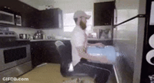 A Man With A Beard Is Sitting In A Chair In A Kitchen Holding A Bucket . GIF