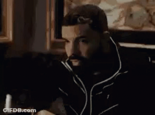 A Man With A Beard Is Sitting In A Chair In Front Of A Painting In A Room . GIF
