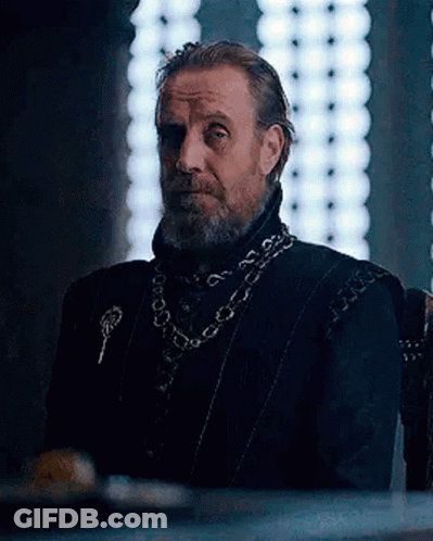 A Man With A Beard Is Sitting In A Chair In Front Of A Window Wearing A Chain Around His Neck . GIF