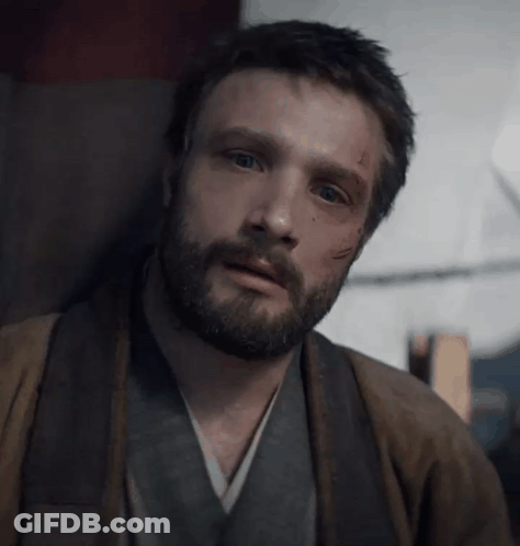 A Man With A Beard Is Sitting In A Chair Looking At The Camera . GIF