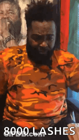 A Man With A Beard Is Sitting In A Chair With His Arms Outstretched And A Camouflage Shirt On . GIF