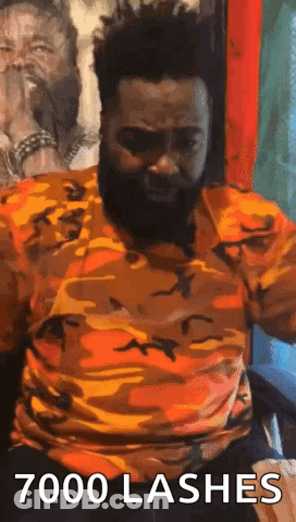 A Man With A Beard Is Sitting In A Chair With His Arms Outstretched And A Camouflage Shirt On . GIF