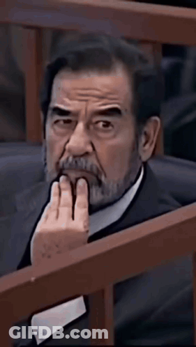 A Man With A Beard Is Sitting In A Chair With His Hand On His Chin And Looking At The Camera . GIF