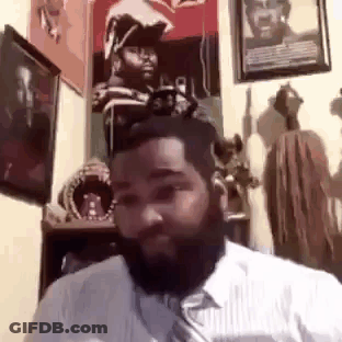 A Man With A Beard Is Sitting In A Room With A Lot Of Paintings On The Wall . GIF