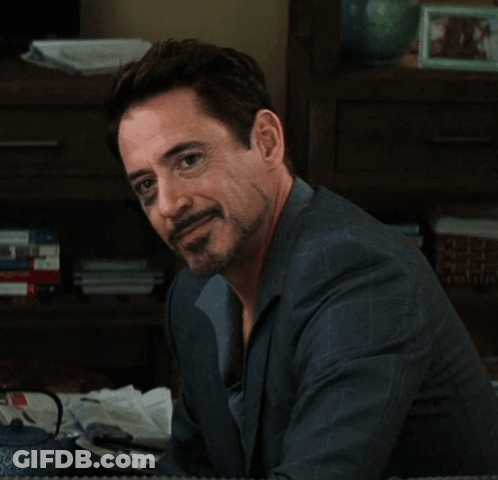 A Man With A Beard Is Sitting In Front Of A Bookshelf And Looking At The Camera GIF
