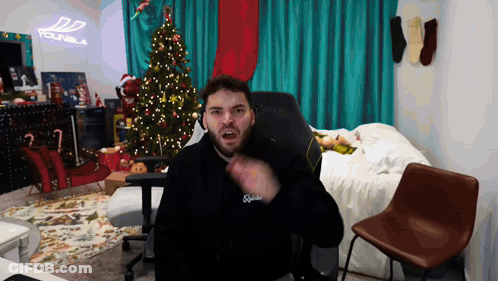 A Man With A Beard Is Sitting In Front Of A Christmas Tree And A Sign That Says Younbla GIF