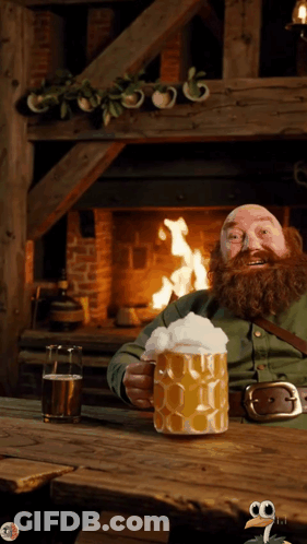 A Man With A Beard Is Sitting In Front Of A Fireplace Holding A Large Mug Of Beer GIF
