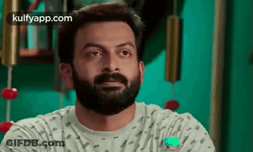 A Man With A Beard Is Sitting In Front Of A Green Wall And Looking At The Camera . GIF