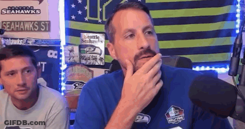 A Man With A Beard Is Sitting In Front Of A Microphone And Covering His Mouth With His Hand . GIF
