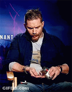 A Man With A Beard Is Sitting In Front Of A Sign That Says Nant . GIF