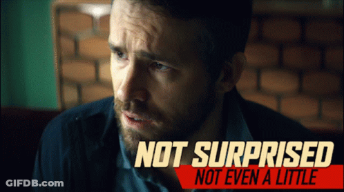 A Man With A Beard Is Sitting In Front Of A Sign That Says Not Surprised GIF