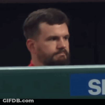 A Man With A Beard Is Sitting In Front Of A Sign That Says S. GIF