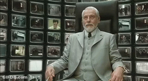 A Man With A Beard Is Sitting In Front Of A Wall Of Televisions GIF