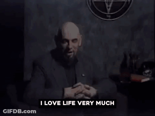 A Man With A Beard Is Sitting In Front Of A Wall That Says I Love Life Very Much GIF