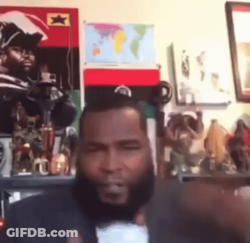 A Man With A Beard Is Sitting In Front Of A Wall With A Map Of The World . GIF