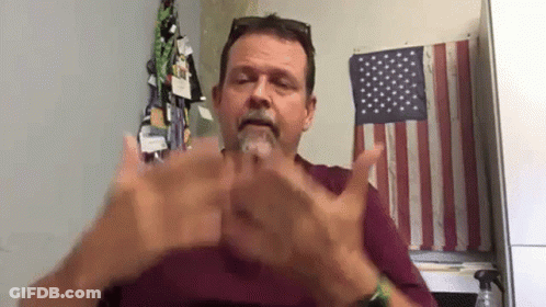A Man With A Beard Is Sitting In Front Of An American Flag And Making A Hand Gesture . GIF