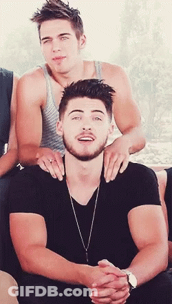 A Man With A Beard Is Sitting Next To Another Man With His Hands On His Shoulders GIF