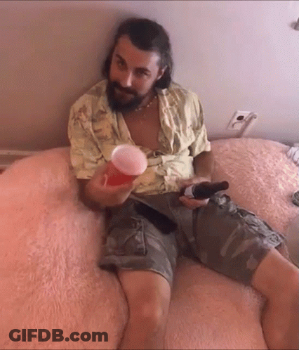A Man With A Beard Is Sitting On A Bed Holding A Beer And A Cup GIF