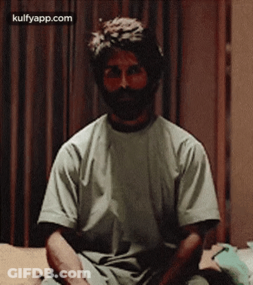 A Man With A Beard Is Sitting On A Bed Wearing A T-Shirt . GIF