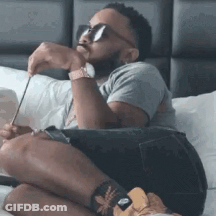 A Man With A Beard Is Sitting On A Bed Wearing Sunglasses And A Watch . GIF