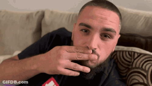 A Man With A Beard Is Sitting On A Couch And Covering His Mouth With His Hand GIF