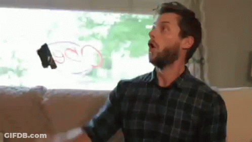 A Man With A Beard Is Sitting On A Couch In Front Of A Window Holding A Remote Control In His Hand . GIF