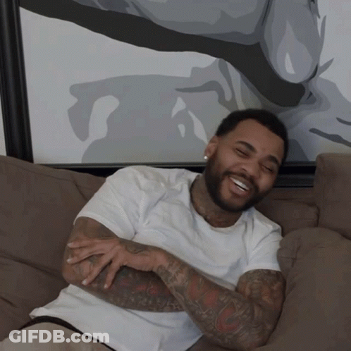 A Man With A Beard Is Sitting On A Couch Smiling GIF