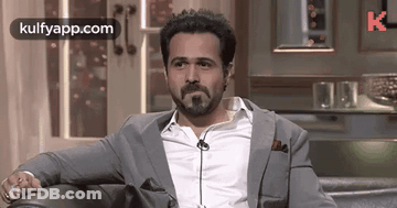 A Man With A Beard Is Sitting On A Couch Wearing A Suit And Tie . GIF