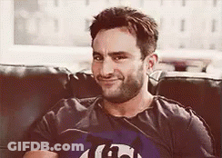 A Man With A Beard Is Sitting On A Couch Wearing A T-Shirt That Says G On It . GIF