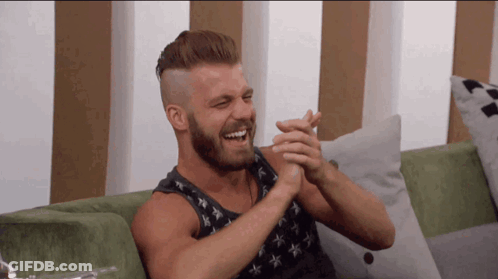 A Man With A Beard Is Sitting On A Green Couch And Clapping His Hands GIF