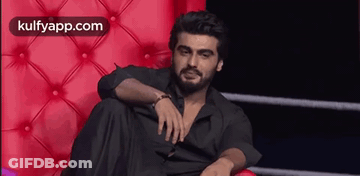 A Man With A Beard Is Sitting On A Red Couch With His Legs Crossed . GIF