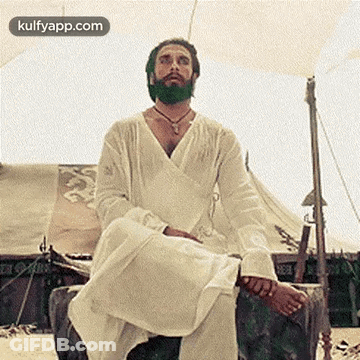 A Man With A Beard Is Sitting On A Rock In Front Of A Tent On The Beach . GIF