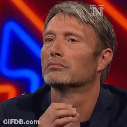 A Man With A Beard Is Sitting On A Stage With His Hands Folded In Front Of His Face . GIF