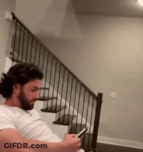 A Man With A Beard Is Sitting On A Staircase Looking At His Phone . GIF